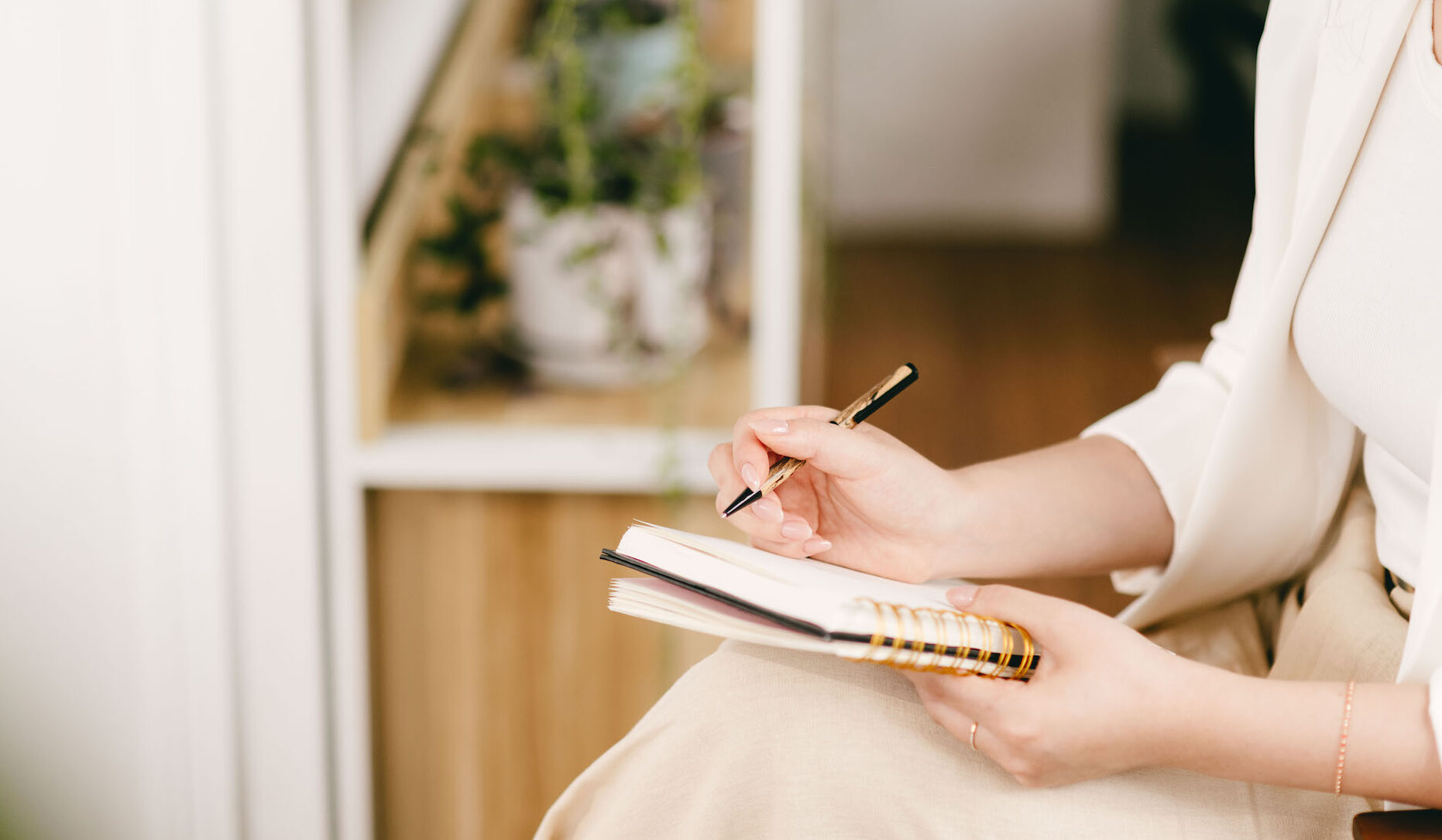 Woman's left hand holding a notebook and a pen in her right hand with plants on a shelf in the background.