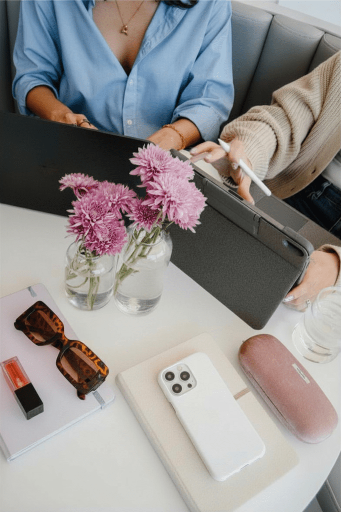 A faceless image of two women interacting with their tablets on a coffee table with a glass vase and pink flowers.